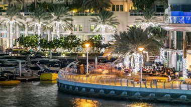 Yürüyen insanlarla dolaş ve Dubai Marina Kanalı üzerinde trafiği olan köprü gece vakti, BAE. Palmiyeler, tekneler ve kulelerle rıhtımın manzarası. Yapay kanallı bölge