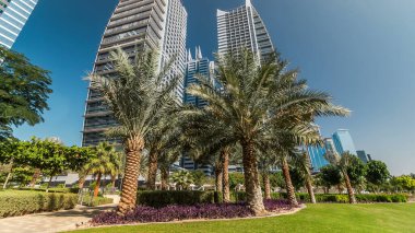 Parktaki gökdelenlerin palmiyeli görüntüsü. Dubai, BAE 'deki Jumeirah Gölü Kuleleri' nde camla kaplı konut binaları. Çimlerden görünüm