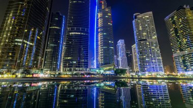Rıhtımdaki ışıklandırılmış gökdelenlerin gece görüş açısını hızlandırın. Dubai, BAE 'deki Jumeirah Gölü Kuleleri' ndeki konut binaları. Rıhtımdan suya yansıyan görüntüyü göster