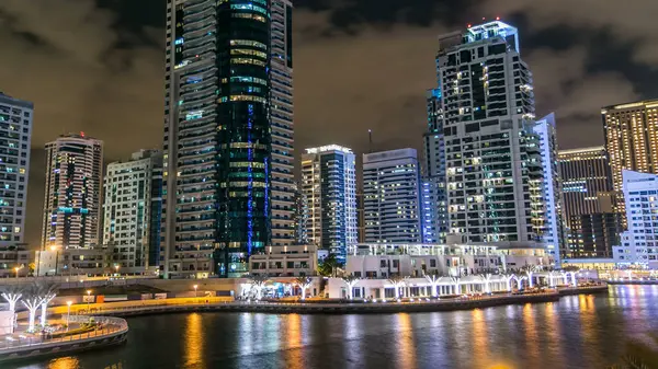 Dubai Marina promenade with boats, palms and modern towers reflected in water from waterfront in Dubai night, United Arab Emirates. District with artificial canal and illuminated windows in skyscraper