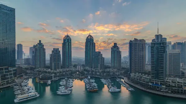 View of modern skyscrapers shining in sunrise lights timelapse in Dubai Marina with yachts in Dubai, UAE. Rays of lights behind towers. Orange clouds on the sky