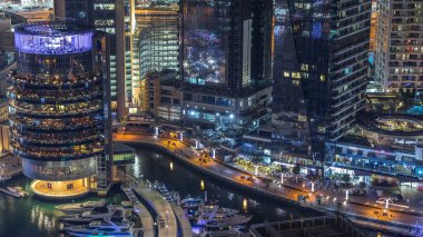Dubai Marina hava saatinin gece aydınlanması, BAE. Modern gökdelenler ve konut binaları. İnsanlar gezinti güvertesinde yürüyor. Yapay kanal şehrindeki alışveriş merkezinin yakınındaki yatlar ve tekneler.