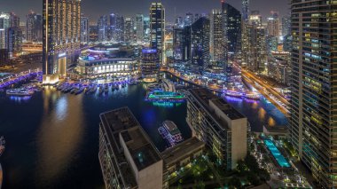 Dubai Marina hava saatinin gece aydınlanması, BAE. Modern gökdelenler ve konut binaları. Yollarda trafik vardı. Yapay kanal şehrindeki alışveriş merkezinin yakınındaki yatlar ve tekneler.