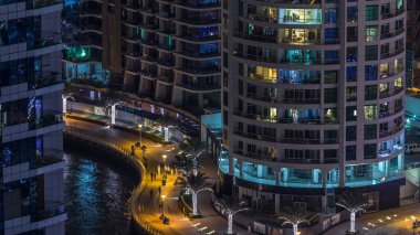 Dubai Marina hava saatinin gece aydınlanması, BAE. Modern gökdelenler ve konut binaları. İnsanlar gezinti güvertesinde yürüyor. Yapay kanal şehrindeki alışveriş merkezinin yakınındaki yatlar ve tekneler.