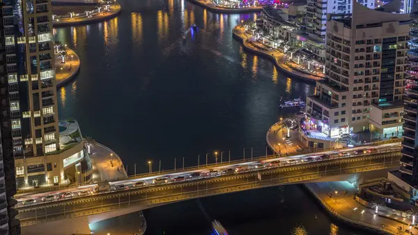 Water canal and promenade on Dubai Marina skyline at night timelapse. Residential towers with lighting and illumination. Floating yachts and boats with traffic on a bridge near skyscrapers