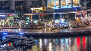 Lüks Dubai Marina kanalı, oradan geçen tekneler ve restoranlarla dolu bir gezinti yeri ve kafeterya gece zaman çizelgesi, yukarıdan aydınlık gökdelenleri olan bir manzara. Dubai, Birleşik Arap Emirlikleri