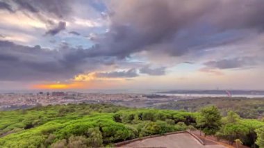 Panoramic sunrise view over Lisbon and Almada from a viewpoint in Monsanto above green trees at morning timelapse. Aerial top overview with colorful clouds and rays of light