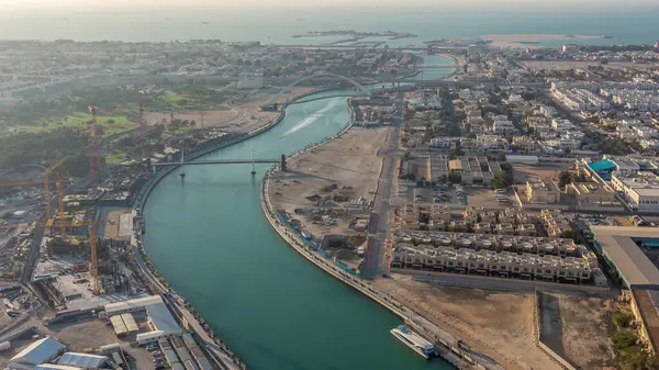 Dubai water canal with footbridge during sunset aerial timelapse from Downtown skyscrapers rooftop. Floating boats and construction site with cranes