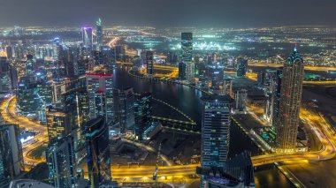 Dubai gece zaman atlamalı iş bölümü kuleleri panoramik havadan görünümü. Yapım aşamasındaki bazı ışıklı gökdelenlerin, kanalın ve yeni kulelerin çatı görünümü.