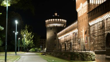 Sforza Kalesi ve kulesinin ana girişi Castello Sforzesco gece vakti aydınlatılmış, Milano, İtalya. Bayraklı yan görünüm