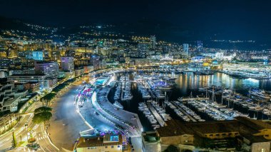Monte Carlo 'lu Panorama, Monaco köyündeki gözlem güvertesinden Herkül Limanı ile birlikte. Aydınlatmalı binalar ve liman manzaralı yatlar. Yolda trafik vardı.