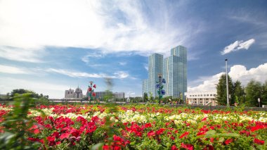 Flowerbed with red and yellow flowers on a waterfront near river timelapse hyperlapse on the background of blue sky with clouds and green towers. Nur-Sultan city, Kazakhstan. Sunny day. clipart