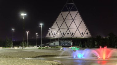 Gece, Barış ve Uzlaşma Sarayı 'nın yanında renkli fıskiyelerle dolu bir meydan. Kazakistan' ın Nur-Sultan şehrinde zaman kavramını aydınlatan bir bina.