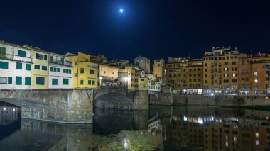 Famous Ponte Vecchio bridge timelapse hyperlapse over the Arno river in Florence, Italy, lit up at night. Full moon rising. Reflection on water. Old houses on the side clipart