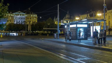 New modern trams of Croatian capital Zagreb night timelapse near railway station. People at tram stop at evening time. CROATIA