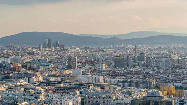 stock image Aerial panoramic view of Vienna city with skyscrapers, historic buildings and mountains on a background timelapse in Austria. Evening skyline before sunset from Danube Tower viewpoint.