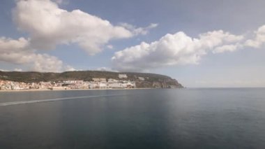 Panorama, Portekiz 'deki Sesimbra Town ve Port Timelapse manzarasını gösteriyor. İskeledeki deniz fenerinden kayıklar, evler ve plajlarla dolu bir gökyüzü manzarası. Setubal bölgesinde bir tatil köyü. Güzel bulutlar