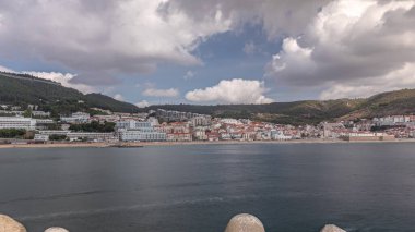 Panorama showing view of Sesimbra Town and Port timelapse, Portugal. Skyline landscape with boats, houses and beach from lighthouse on a pier. Resort in Setubal district clipart
