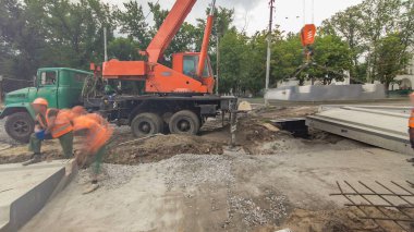 Yol inşaatında vinçle beton plakalar yerleştiriliyor. Zaman ayarlı hiperhızlandırma. Şapkalı ve üniformalı endüstriyel işçiler. Şehir caddesindeki tramvay raylarının yeniden inşası devam ediyor.