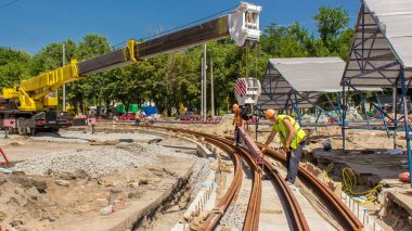 Sarı inşaat teleskopik mobil vinç, tramvay raylarını beton plakalara taşıyor. Şapkalı ve üniformalı endüstriyel işçiler. Şehir caddesindeki tramvay raylarının yeniden inşası