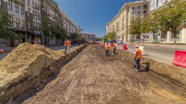 Turuncu üniformalı işçiler yol inşaatında toprak kazıyor ve zaman aşımına uğruyor. Arka planda endüstriyel sarı kazıcı. Kavşaktaki tramvay raylarının yeniden yapılandırılmasının görüntüsü