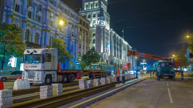 Turuncu yapım teleskopik mobil vinç kamyon gecesinden tramvay raylarını boşaltıyor. Şapkalı ve üniformalı endüstriyel işçiler. Şehir caddesindeki tramvay raylarının yeniden inşası