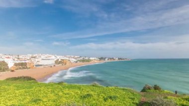 Panorama Albufeira Timelapse şehrinde geniş kumlu sahil ve Atlantik okyanusunu gösteriyor. Uçurumun tepesindeki beyaz evler. Yeşil çimlerin üzerinde sarı çiçeklerle yukarıdan hava manzarası. Algarve, Portekiz