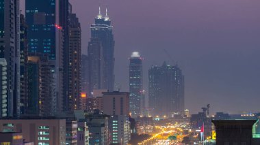 Dubai Kuleleri 'nin şehir merkezinden gece gündüz geçiş zamanı. Gün batımından sonra Şeyh Zayed 'in gökdelenli hava manzarası. Yoldaki trafik ve çok katlı otopark.