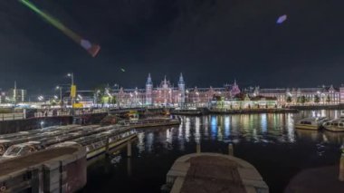 Panorama aydınlatılmış Central Railway İstasyonu gece saat çizelgesini gösteriyor. Hollanda, Amsterdam 'daki tren istasyonunun önündeki bir iskeleye, tramvaylara ve kanallara demirlemiş tur tekneleri