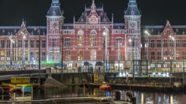 Aydınlatılmış Central Railway İstasyonu gece zaman çizelgesi. Hollanda, Amsterdam 'daki tren istasyonunun önündeki bir iskeleye, tramvaylara ve kanallara demirlemiş tur tekneleri