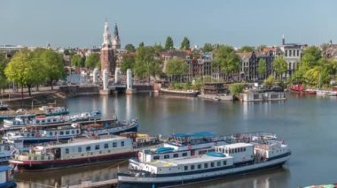 Amsterdam 'ın şehir panoramasının önünde Montelbaanstoren kulesi ve kanal boyunca uzanan şehir manzarası yer almaktadır. Eski kentin rıhtımında tarihi Hollanda evleri, mavnalar ve yüzen evler