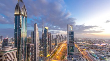Dubai şehir mimarisi günden geceye geçiş zamanı. Şeyh Zayed yolu üzerindeki aydınlık gökdelenler ve trafik manzaralı üst hava manzarası. Güzel bulutlu gökyüzü