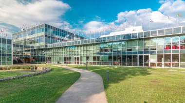 İş bölgesindeki modern düşük katlı cam ofis binası ve iç avlu zaman aralığında hızlandırılmış yol olan yeşil çimenler. Çatıda pek çok farklı ülkenin bayrakları