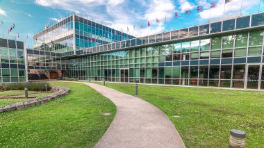 İş bölgesindeki modern düşük katlı cam ofis binası ve iç bahçedeki yeşil çimenlik zaman ayarlı hiperhızlandırıcı. Çatıda pek çok farklı ülkenin bayrakları var. Mavi bulutlu gökyüzü