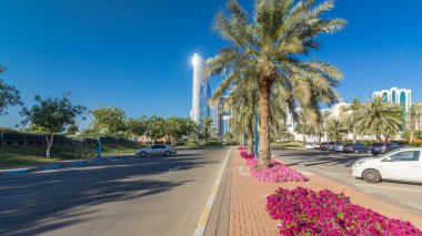 Abu Dabi 'nin sahil şeridi boyunca uzanan Corniche Bulvarı plajı parkında arka planda gökdelenler var. Aile parkından görüntü. Palmiyeler ve çiçekler yan yana. Güneşli bir günde mavi gökyüzü
