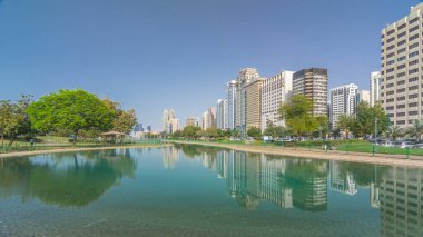Abu Dabi 'nin sahil şeridi boyunca uzanan Corniche Bulvarı plajı parkında arka planda gökdelenler var. Lake Park 'tan yansıtmalı görüntü. Bir yanda ağaçlar