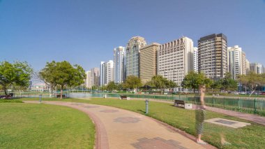 Abu Dabi 'nin sahil şeridi boyunca uzanan Corniche Bulvarı plajı parkında arka planda gökdelenler var. Lake Park 'ın manzarası. Bir tarafta ağaçlar. Güneşli bir günde mavi gökyüzü