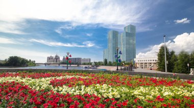 Nehir kıyısında kırmızı ve sarı çiçeklerle süslü bir çiçek tarhı. Zaman atlaması, mavi gökyüzünün arka planında bulutlar ve yeşil kuleler. Nur-Sultan şehri, Kazakistan. Güneşli bir gün.