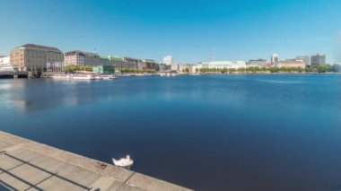 Panorama, Almanya, Hamburg 'da Binnenalster zaman çizelgesini tekne istasyonuyla gösteriyor. Alster Gölü 'nün iç sularına yansıyan tarihi binalar mavi gökyüzünün altında. İskelede kuğularla manzaralı şehir manzarası