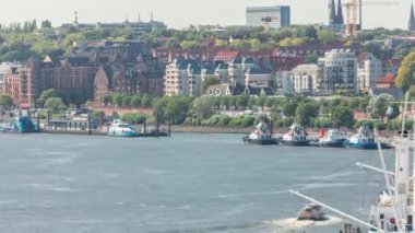 Almanya 'nın Hamburg kentinde Elbe nehir kenarının hava zaman çizelgesi. İskeleye yakın tekneler, marina ve geçen gemilerle manzara. İnşaat alanında vinçleri olan evler. Elbe Nehri kıyısındaki canlı şehir manzarası