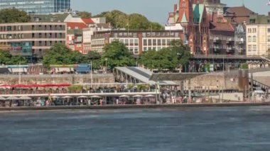 Hamburg limanına giriş ve çıkış yapan bir feribotun rıhtımdaki yolcuları ve merdivenleri gösteren zaman çizelgesi. Arka planda Elbe nehri boyunca tarihi ve modern binalar var. Almanya