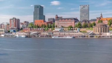 Hamburg limanına giriş ve çıkış yapan bir feribotun zaman çizelgesi. İskelede yolcuları var ve merdivenlerde ilerliyor. Arkaplanda Elbe nehri boyunca tarihi ve modern binalar var. Almanya