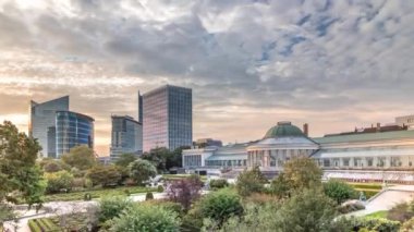 Le Botanique 'nin Brüksel, Belçika' daki havadan çekilmiş zaman çizelgesi. Etrafı bahçeler ve kentsel gökdelenlerle çevrili. Gün batımında kubbe ve yürüyen insanlar canlı şehir manzarasında turuncu bulutlarla