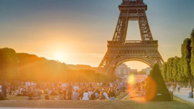 Eiffel Tower seen from Champ de Mars during sunset timelapse, Paris, France. People sitting on lawn. Summer evening with clear sky. Orange sky and rays of light clipart