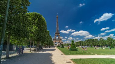Paris 'teki Champs de Mars' taki Eyfel Kulesi. Zaman atlaması, Fransa. Yazın mavi bulutlu gökyüzü Ağaçlı yeşil çimenler ve etrafta yürüyen insanlar.