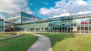 İş bölgesindeki modern düşük katlı cam ofis binası ve iç avlu zaman aralığında hızlandırılmış yol olan yeşil çimenler. Çatıda pek çok farklı ülkenin bayrakları