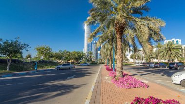 Abu Dabi 'nin sahil şeridi boyunca uzanan Corniche Bulvarı plajı parkında arka planda gökdelenler var. Aile parkından görüntü. Palmiyeler ve çiçekler yan yana. Güneşli bir günde mavi gökyüzü