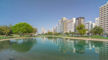 Abu Dabi 'nin sahil şeridi boyunca uzanan Corniche Bulvarı plajı parkında arka planda gökdelenler var. Lake Park 'tan yansıtmalı görüntü. Bir yanda ağaçlar