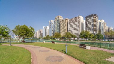 Abu Dabi 'nin sahil şeridi boyunca uzanan Corniche Bulvarı plajı parkında arka planda gökdelenler var. Lake Park 'ın manzarası. Bir tarafta ağaçlar. Güneşli bir günde mavi gökyüzü