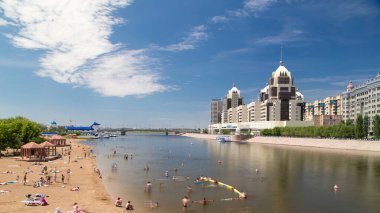 İnsanlar, Astana, Kazakistan 'daki köprüden ve ufuk çizgisinden gelen yapay bir plaj zaman atlaması boyunca bir nehirde banyo yapıyorlar. Nur-Sultan şehri